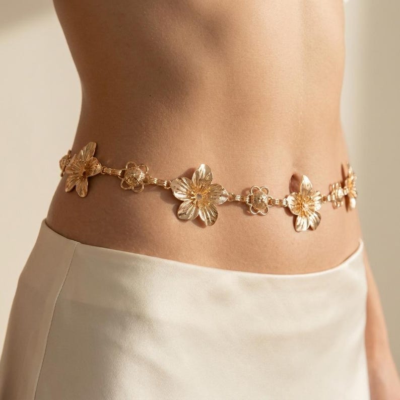 May include: A gold-colored waist chain featuring a floral design. The chain is composed of multiple flower-shaped links connected by smaller, decorative elements. The belt is worn over a white skirt, creating a stylish accessory.