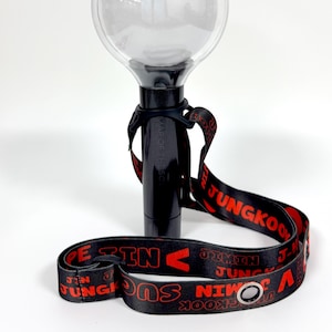 May include: Black and clear concert light stick with a lanyard. The lanyard is black with red text that reads "JUNGKOOK" and other names. The light stick has a clear, round top and a black handle.