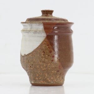 May include: A ceramic jar with a lid, featuring a textured, speckled brown base that transitions into a smooth, glossy white and dark reddish-brown glaze on the upper portion. The vessel has a slightly flared rim and a small, knobbed lid.