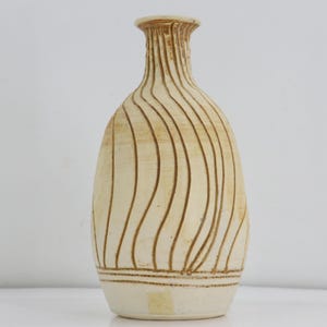 May include: A cream-colored ceramic vase with a narrow neck and a rounded body. The surface features vertical, wavy brown lines that extend from the neck down to a horizontal band near the base. A small rectangular sticker is visible near the bottom.