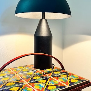 May include: A modern black table lamp with a teal dome-shaped shade. The lamp sits on a small table with a colorful, patterned tile top. The table has a red metal frame and a curved handle.