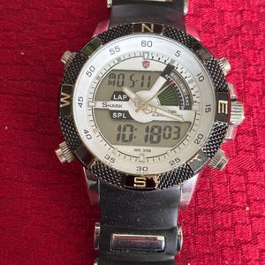 K&ouml;nnte beinhalten: Eine schwarze und silberne digitale Shark-Uhr mit wei&szlig;em Zifferblatt. Die Uhr zeigt die Uhrzeit und das Datum an, mit dem Text "SHARK" und "SPL" sichtbar. Die Uhr hat ein schwarzes Kautschukarmband und eine drehbare L&uuml;nette.