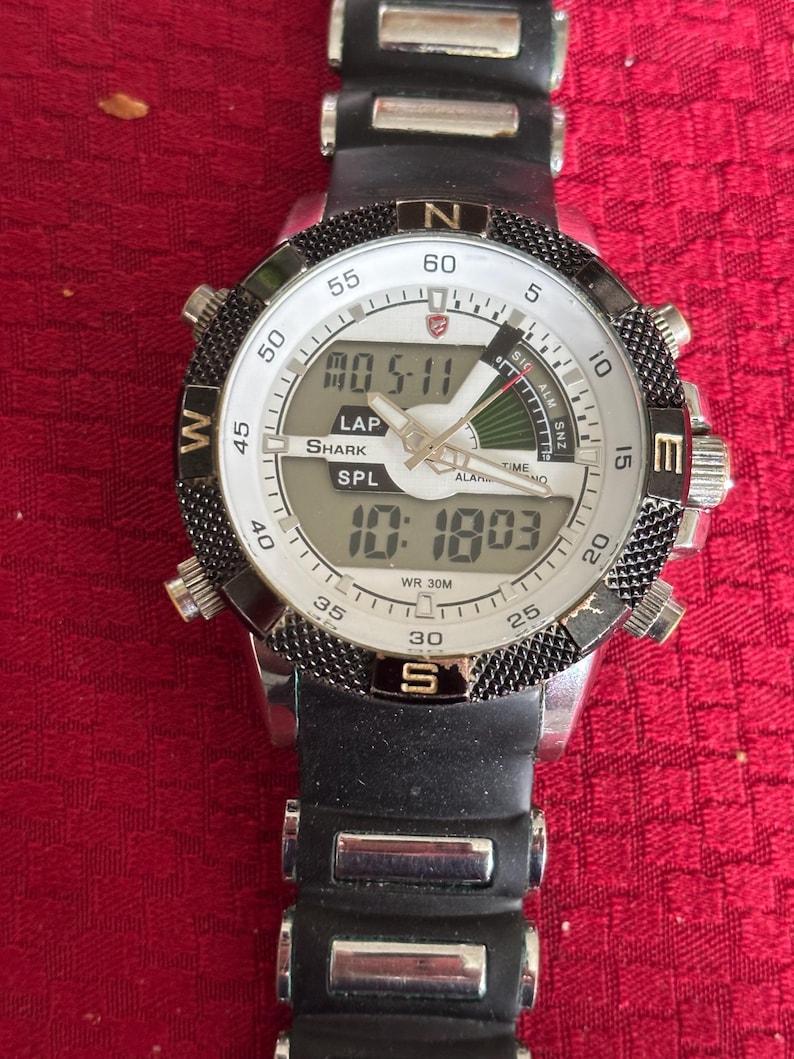 K&ouml;nnte beinhalten: Eine schwarze und silberne digitale Shark-Uhr mit wei&szlig;em Zifferblatt. Die Uhr zeigt die Uhrzeit und das Datum an, mit dem Text "SHARK" und "SPL" sichtbar. Die Uhr hat ein schwarzes Kautschukarmband und eine drehbare L&uuml;nette.