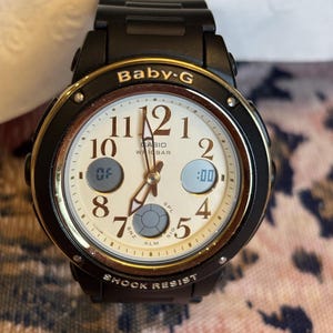 Op de afbeelding: Zwarte Baby-G horloge met een crèmekleurige wijzerplaat en gouden accenten. Het horloge heeft een digitale display en analoge wijzers. De wijzerplaat toont de tekst "Baby-G", "CASIO" en "SHOCK RESIST".