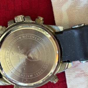 K&ouml;nnte beinhalten: Nahaufnahme der R&uuml;ckseite einer silberfarbenen Uhr mit schwarzem Kautschukarmband. Das Zifferblatt der Uhr tr&auml;gt die Aufschrift "PRECISION MOVEMENT" und "WATER RESISTANT 30M". Die Uhr tr&auml;gt auch die Aufschrift "SHARK SPORT WATCH" und "STAINLESS STEEL".