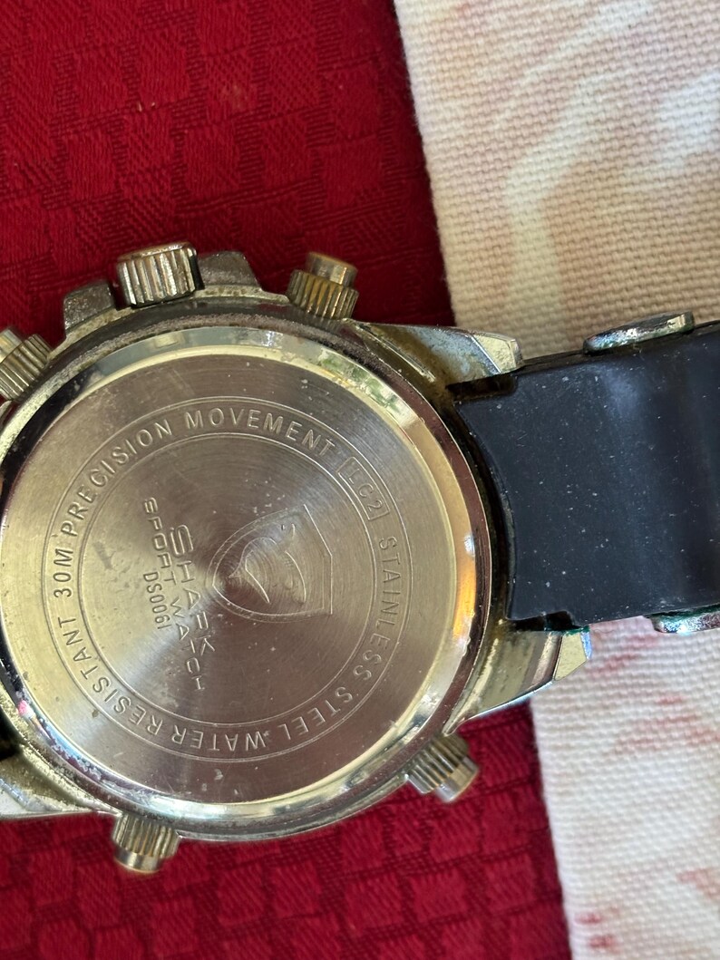 K&ouml;nnte beinhalten: Nahaufnahme der R&uuml;ckseite einer silberfarbenen Uhr mit schwarzem Kautschukarmband. Das Zifferblatt der Uhr tr&auml;gt die Aufschrift "PRECISION MOVEMENT" und "WATER RESISTANT 30M". Die Uhr tr&auml;gt auch die Aufschrift "SHARK SPORT WATCH" und "STAINLESS STEEL".