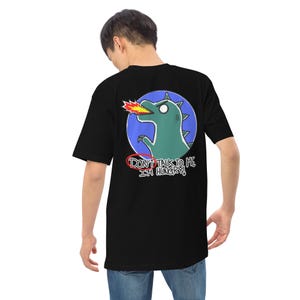 Puede incluir: Camiseta negra con un dinosaurio verde de dibujos animados que escupe fuego, sobre un círculo azul. El texto "DON'T TALK TO ME I'M HUNGRY!" está impreso debajo. Prenda informal.