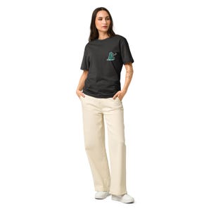 Puede incluir: Una persona con una camiseta negra con un pequeño diseño bordado en el pecho, combinada con pantalones color crema y zapatillas blancas. La camiseta presenta un diseño verde y azul. Los pantalones tienen un corte de pierna ancha.