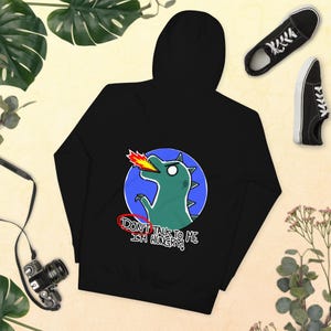 Puede incluir: Sudadera con capucha negra con un diseño de dinosaurio de dibujos animados. El dinosaurio verde escupe fuego por la boca, sobre un círculo azul. El texto "DON'T TALK TO ME I'M HUNGRY!" está debajo. Zapatillas negras y una cámara en el fondo.