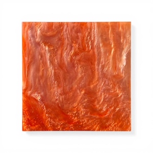 Könnte beinhalten: Quadratischer, orangefarbener Dekorationsblock mit einem wirbelnden, marmorierten Muster. Die Oberfläche hat ein glänzendes Finish mit dezenten Glitzerakzenten, wodurch ein schimmernder Effekt entsteht. Der Block ist vor einem weißen Hintergrund platziert.