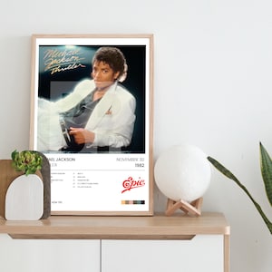 Puede incluir: Un póster enmarcado con Michael Jackson con un traje blanco, con el título "Thriller". El póster incluye la fecha de lanzamiento del álbum y el logotipo de Epic Records. La imagen se muestra en un mueble de madera, con un jarrón decorativo y una lámpara.