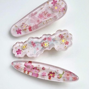 以下が含まれることがあります： ピンクの桜の花のデザインが施された、3つの樹脂製ヘアクリップ。クリップは、バレッタと2つのスナップクリップなど、さまざまな形状をしています。金箔がピンクの花をアクセントにしています。白い背景に配置されています。