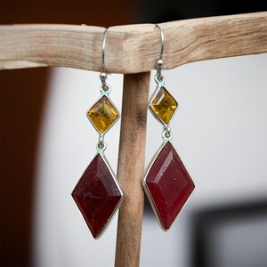May include: A pair of drop earrings featuring silver-toned metal hooks and settings. Each earring consists of a small, faceted, amber-coloured diamond-shaped stone positioned above a larger, faceted, deep red diamond-shaped stone.