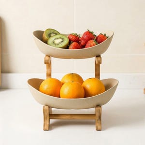 May include: A two-tiered fruit stand featuring two beige, oval-shaped bowls supported by a light brown bamboo frame. The top bowl contains sliced green kiwi and red strawberries, while the bottom bowl holds three whole bright orange citrus fruits.