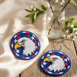May include: A pair of patriotic earrings featuring a detailed embroidered eagle head design. The earrings have a blue background with white stars and red accents, and a silver-tone hook.