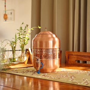 May include: A copper water dispenser with a hammered texture band. The dispenser has a spigot with a blue handle and a lid with a small handle. It sits on a wooden table with a floral runner.