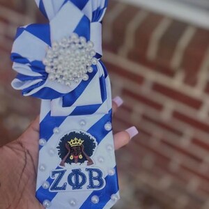 May include: A blue and white striped necktie featuring a large, circular cluster of white faux pearls at the top knot. The lower portion of the tie is decorated with scattered white faux pearls and an embroidered patch of a figure with a crown.