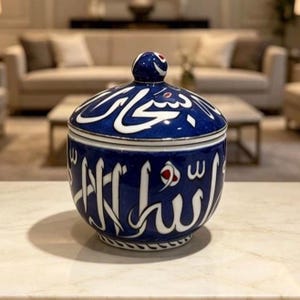 May include: A round ceramic jar with a matching lid, featuring a deep blue glazed finish and prominent white Arabic calligraphy script wrapping around the exterior. The design includes small red accents within the lettering and a knobbed lid.