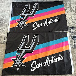 May include: Two black flags featuring the San Antonio Spurs logo. The logo includes a silver spur, a star, and the words "San Antonio" in white. The flags have diagonal stripes in teal, pink, orange, and black.