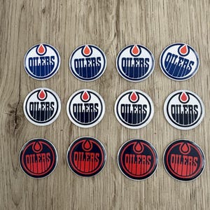 May include: Twelve circular pins featuring the Oilers logo. The pins display the team's logo in blue, white, and orange, with the word "OILERS" in a bold font. The pins are arranged on a wooden surface.