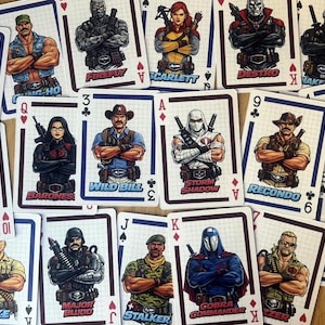 Può includere: Un mazzo di carte da gioco G.I. Joe con vari personaggi come Duke, Cobra Commander e Storm Shadow. Le carte sono disposte su una superficie di legno, con il logo G.I. Joe sul retro. Le carte sono di dimensioni standard.