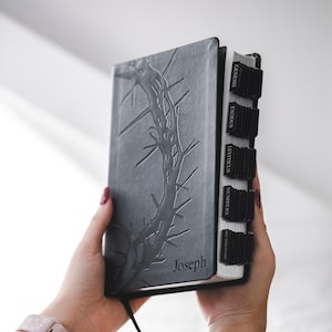 May include: A gray Bible with a thorn design embossed on the cover. The name "Joseph" is printed at the bottom. Black tabs mark the book sections. The Bible is held open, revealing the pages.