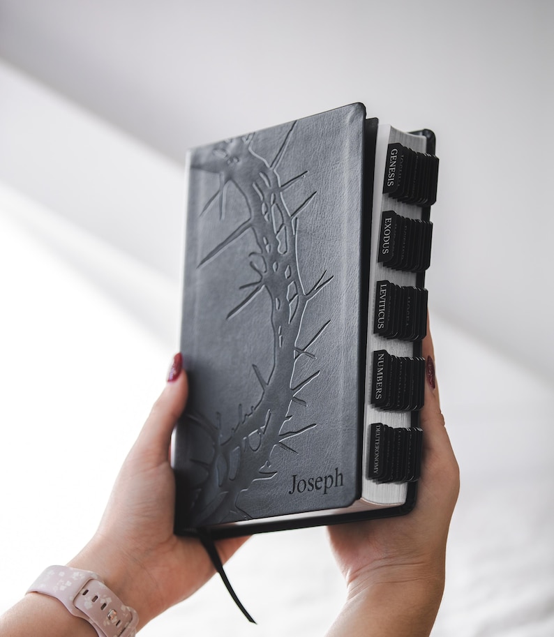 May include: A gray Bible with a thorn design embossed on the cover. The name "Joseph" is printed at the bottom. Black tabs mark the book sections. The Bible is held open, revealing the pages.