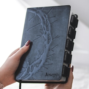 May include: A dark gray Bible with a textured, embossed design and the name "Joseph" on the cover. The book has black tabs labeled with book names and a black ribbon bookmark. The cover design resembles a tree branch.