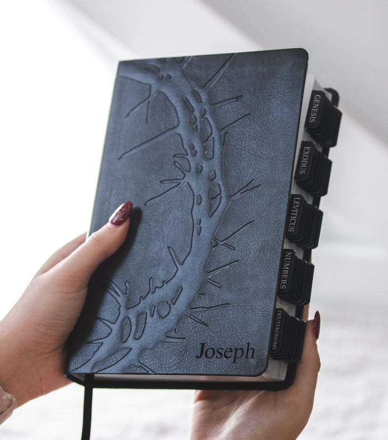 May include: A dark gray Bible with a textured, embossed design and the name "Joseph" on the cover. The book has black tabs labeled with book names and a black ribbon bookmark. The cover design resembles a tree branch.