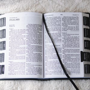 May include: Open Bible with black tabs labeled with book names, including Psalms, Isaiah, and Proverbs. A black ribbon bookmark lies between the pages. The book is open on a white surface.