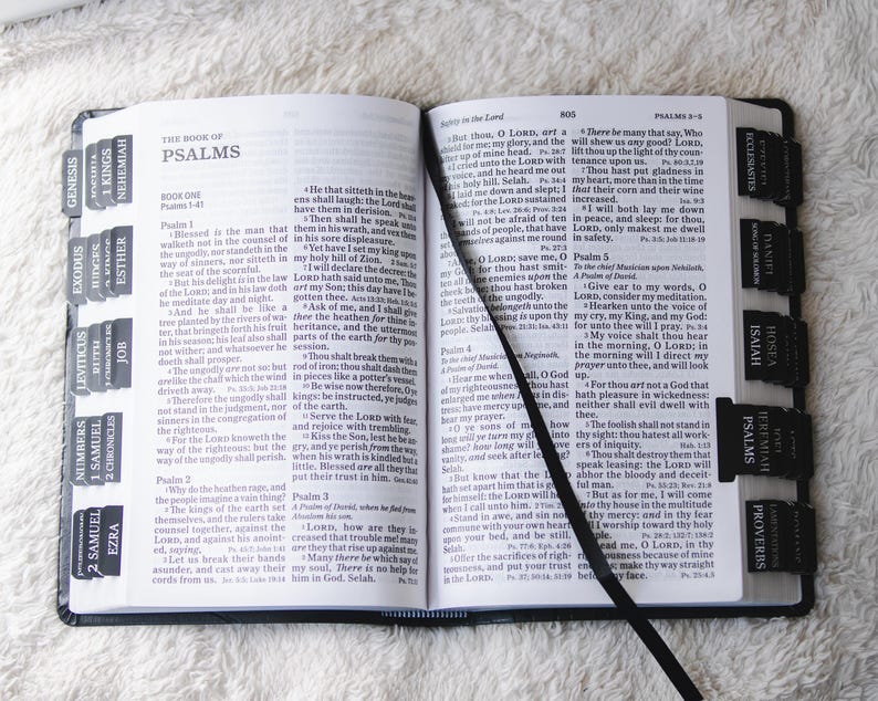 May include: Open Bible with black tabs labeled with book names, including Psalms, Isaiah, and Proverbs. A black ribbon bookmark lies between the pages. The book is open on a white surface.