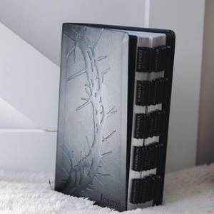 May include: A black Bible with a debossed tree branch design and the name "Joseph" at the bottom. The book has black tabs with the names of the books of the Bible. The book is on a white, fluffy surface.