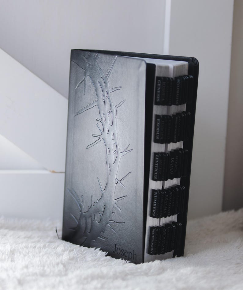 May include: A black Bible with a debossed tree branch design and the name "Joseph" at the bottom. The book has black tabs with the names of the books of the Bible. The book is on a white, fluffy surface.