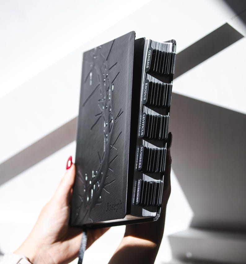May include: A black Bible with embossed botanical design and labeled tabs. The book is held open, revealing the pages and chapter markers. The word "Joseph" is embossed on the cover.