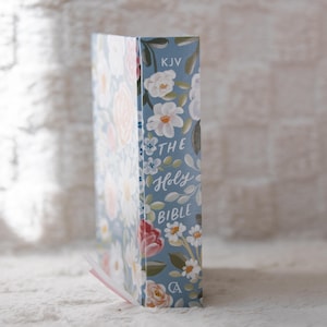 K&ouml;nnte beinhalten: Eine Bibel mit blauem Einband, verziert mit einem Blumenmuster. Das Design umfasst wei&szlig;e, rosa und rote Blumen. Auf dem Buchr&uuml;cken steht "THE Holy BIBLE" und "KJV". Ein rosa Lesezeichen ist sichtbar.