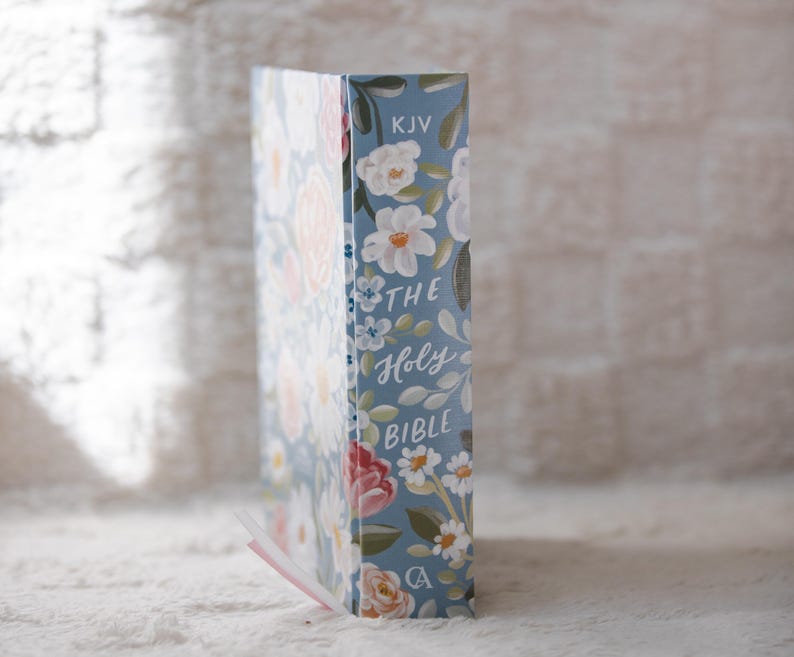 K&ouml;nnte beinhalten: Eine Bibel mit blauem Einband, verziert mit einem Blumenmuster. Das Design umfasst wei&szlig;e, rosa und rote Blumen. Auf dem Buchr&uuml;cken steht "THE Holy BIBLE" und "KJV". Ein rosa Lesezeichen ist sichtbar.