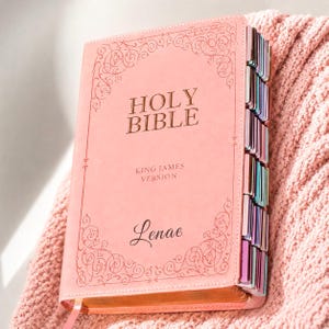 May include: A pink Holy Bible King James Version with gold embossed lettering and decorative scrollwork borders. The cover features the name Lenae in script font. The side of the book includes multi-colored index tabs for navigation.