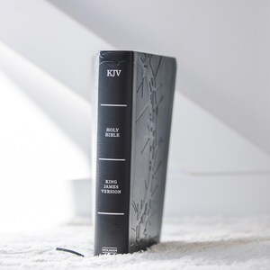 May include: A black leather-bound Holy Bible, King James Version, with embossed detailing. The spine features the text "KJV" and "HOLY BIBLE" in white. The book is standing upright on a white, textured surface.