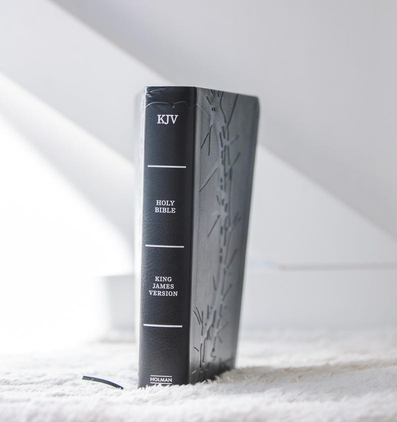 May include: A black leather-bound Holy Bible, King James Version, with embossed detailing. The spine features the text "KJV" and "HOLY BIBLE" in white. The book is standing upright on a white, textured surface.