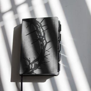 May include: A gray Bible with a black thorn design on the cover. The book has black page dividers and a black ribbon bookmark. The image is lit with natural light, casting shadows.