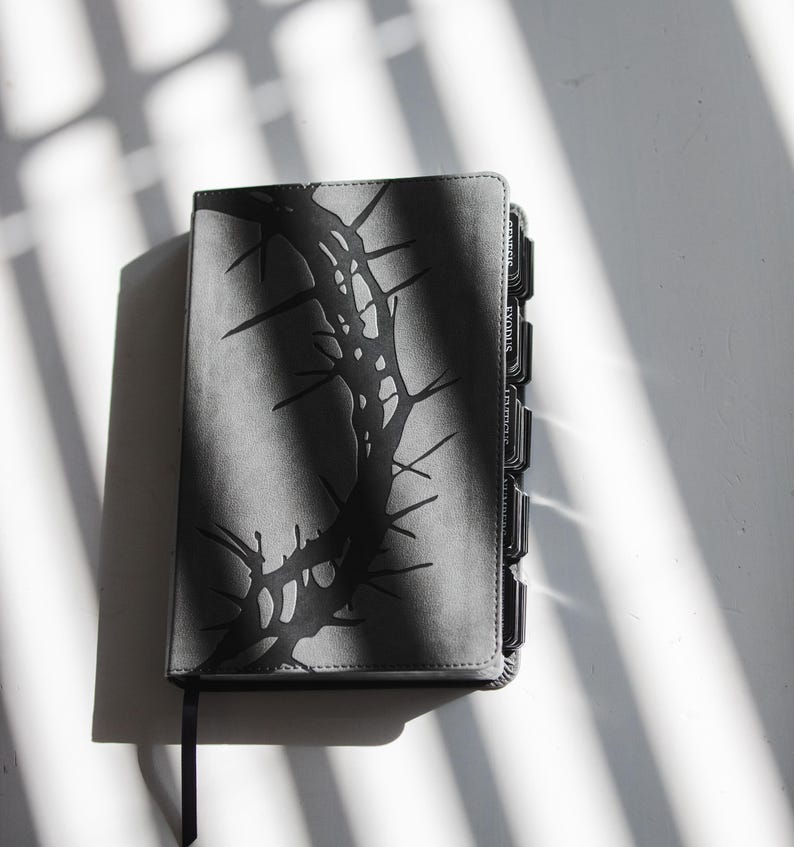 May include: A gray Bible with a black thorn design on the cover. The book has black page dividers and a black ribbon bookmark. The image is lit with natural light, casting shadows.