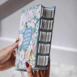 K&ouml;nnte beinhalten: Ein Buch mit hellblauem Einband, verziert mit wei&szlig;en, rosa und orangefarbenen Blumenmustern. Das Buch hat beschriftete Registerkarten und den Text "Do you fix your affection on things above". Das Buch wird offen gehalten.