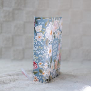 K&ouml;nnte beinhalten: Eine Bibel mit floralem Muster und hellblauem Einband. Der Einband zeigt wei&szlig;e, rosa und orangefarbene Blumen mit gr&uuml;nen Bl&auml;ttern. Auf dem Buchr&uuml;cken steht "THE Holy BIBLE" und "KJV". Ein rosa Lesezeichenband ist sichtbar.