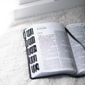May include: Open Bible with black tabs labeled with book names, including Psalms. The book is open to a page of text, with a black ribbon bookmark. The Bible rests on a white, fluffy surface.