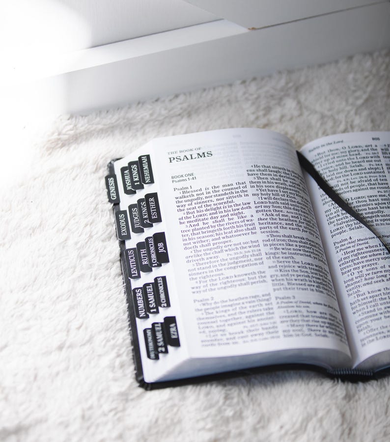 May include: Open Bible with black tabs labeled with book names, including Psalms. The book is open to a page of text, with a black ribbon bookmark. The Bible rests on a white, fluffy surface.