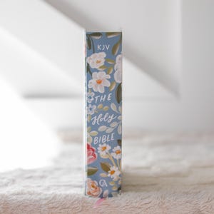 K&ouml;nnte beinhalten: Blauer Buchr&uuml;cken mit floralem Muster, wei&szlig;en und rosa Bl&uuml;ten und gr&uuml;nen Bl&auml;ttern. Der Text "THE Holy BIBLE" ist in wei&szlig;er Schrift gehalten. Die Buchstaben "KJV" befinden sich oben. Ein rosa Lesezeichen ist sichtbar.