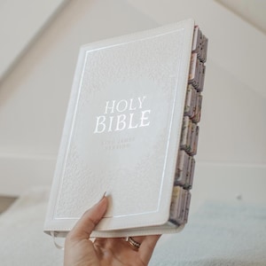 May include: A cream-colored Holy Bible with the text "HOLY BIBLE" and "KING JAMES VERSION" in silver lettering. The book has decorative tabs along the side and a silver border. The Bible is held in a person's hand.