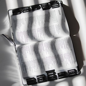 May include: Open Bible with black tabs labeled with book names. The pages are filled with text, and a black ribbon bookmark is visible. The Bible is open to the book of Isaiah.