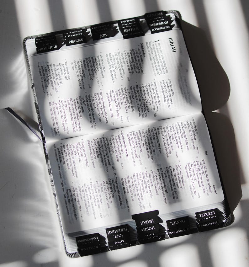 May include: Open Bible with black tabs labeled with book names. The pages are filled with text, and a black ribbon bookmark is visible. The Bible is open to the book of Isaiah.