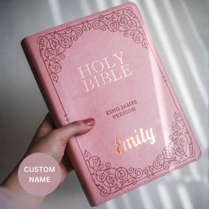 May include: A light pink Holy Bible, King James Version, featuring a decorative border in rose gold foil. The cover is personalized with the name Emily in matching rose gold script lettering. The book has a smooth, leather-like finish.